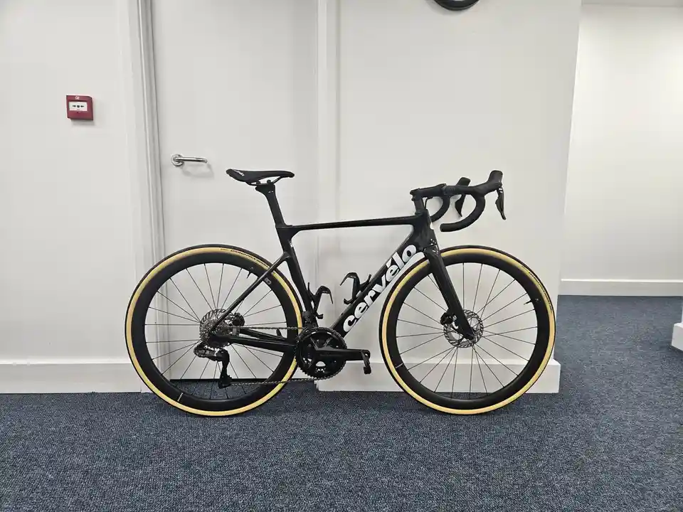 A black Cervélo road bicycle with cream-colored tires is leaning against a white office door on a blue carpeted floor. The…