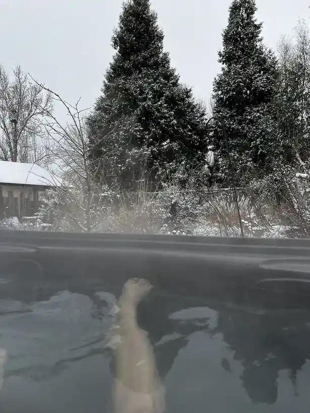 A person's bare foot is visible in steaming hot tub water, with a snowy outdoor scene featuring evergreen trees and a…