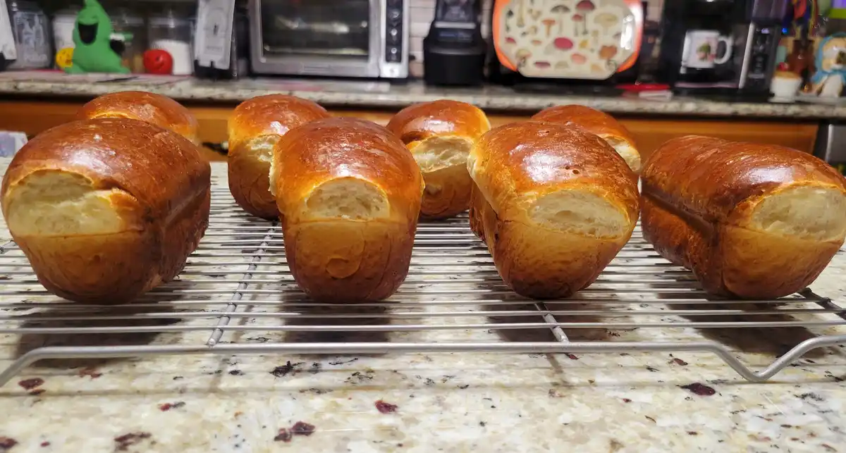 Several golden-brown, freshly baked bread rolls sit on a metal cooling rack on a speckled kitchen countertop. The rolls have…