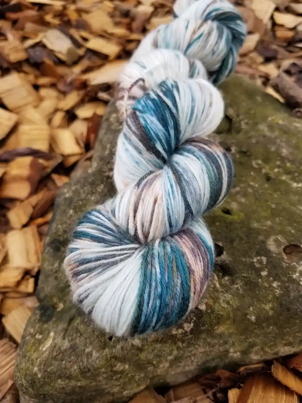 A skein of multicolored yarn with blue, white, and purple hues is coiled on a mossy gray rock, surrounded by wood chips. The…