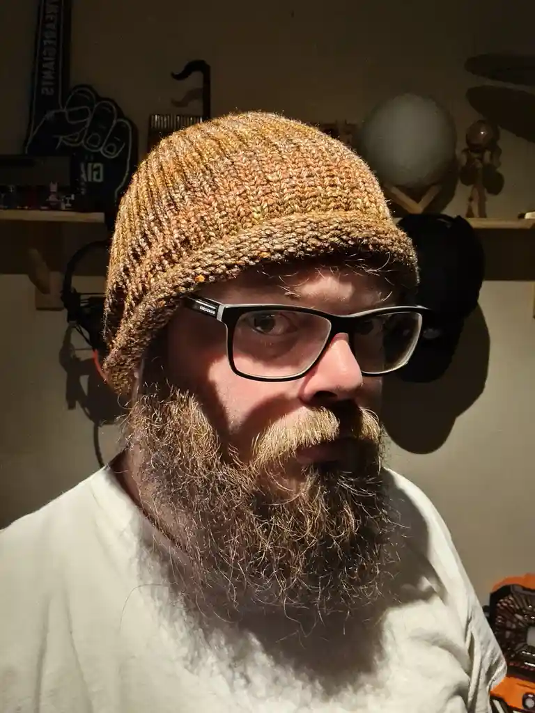 A man with a full beard and glasses wears a brown knitted beanie and looks directly at the camera with a neutral expression…