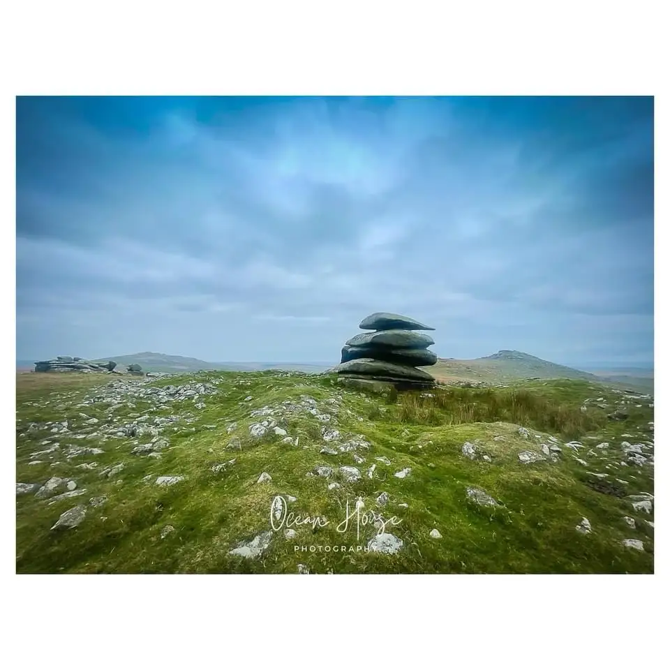 A scenic landscape photo shows a grassy hill dotted with rocks under a cloudy blue sky, featuring a prominent stack of…