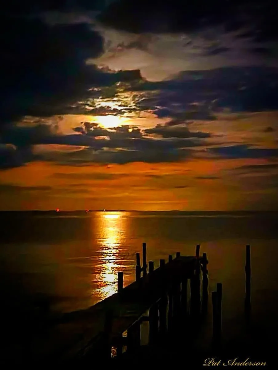 A dramatic sunset over water with a silhouetted wooden pier extending into the frame. The sky is filled with dark clouds…
