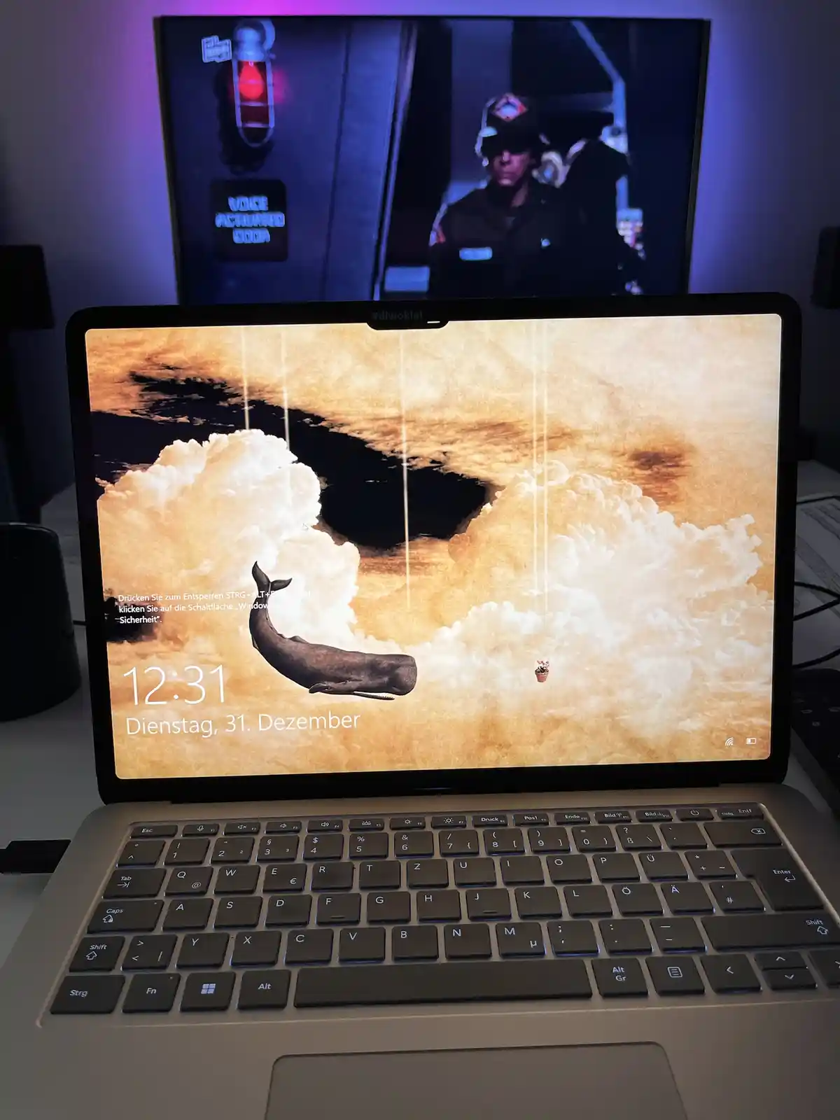 A laptop screen displays a Windows lock screen with a whale wallpaper and German instructions to unlock, while a TV behind…