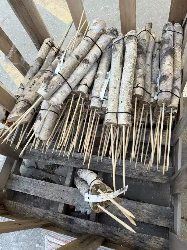 A wooden crate holds numerous birch logs bundled with black ties, each with a long wooden skewer protruding from the center…