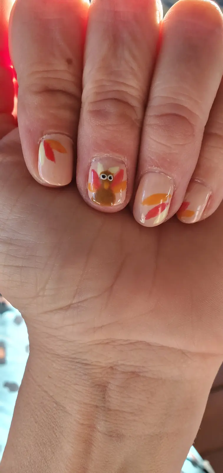 A close-up of a person's hand showing autumn-themed nail art; the middle finger has a cartoon turkey with googly eyes and…