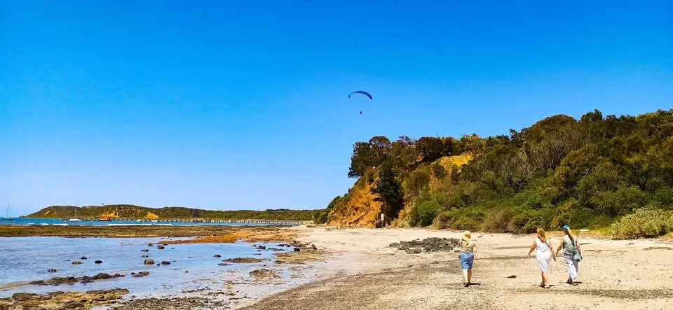 Three people walk along a sunny beach with a paraglider visible in the sky above a cliffside. The overlaid text reads…