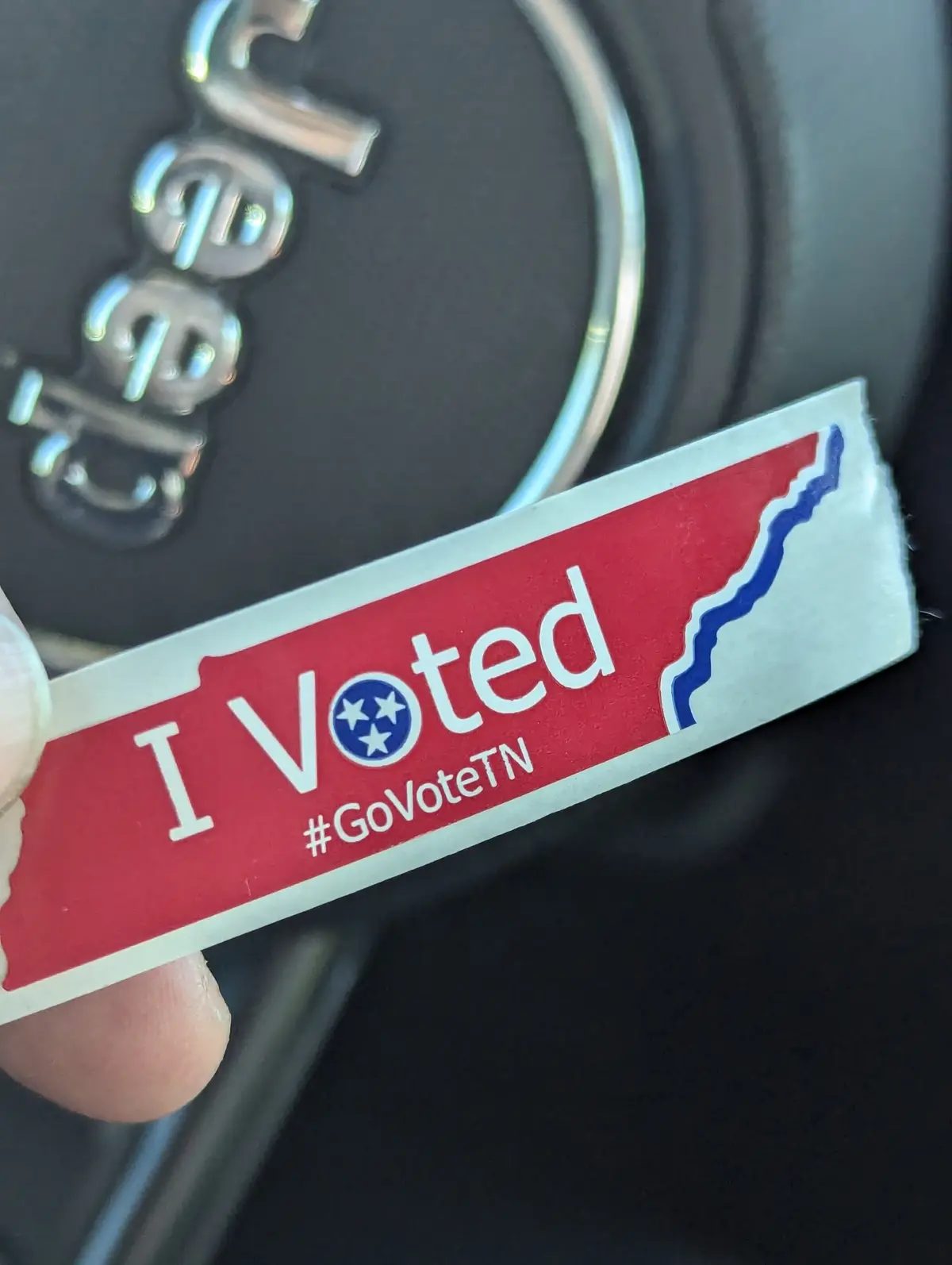 A close-up photo shows a hand holding a red, white, and blue 'I Voted' sticker shaped like the state of Tennessee, featuring…