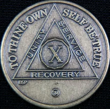 A close-up of a silver-colored X Recovery coin, featuring a large triangle with the words 'TO THINE OWN SELF BE TRUE' around…