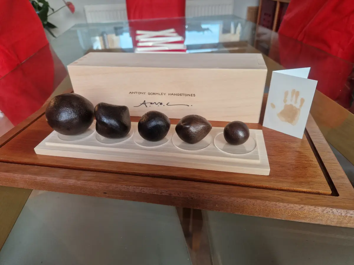 Five dark, hand-shaped stone sculptures are displayed in a row on a wooden base with clear acrylic stands, positioned in…