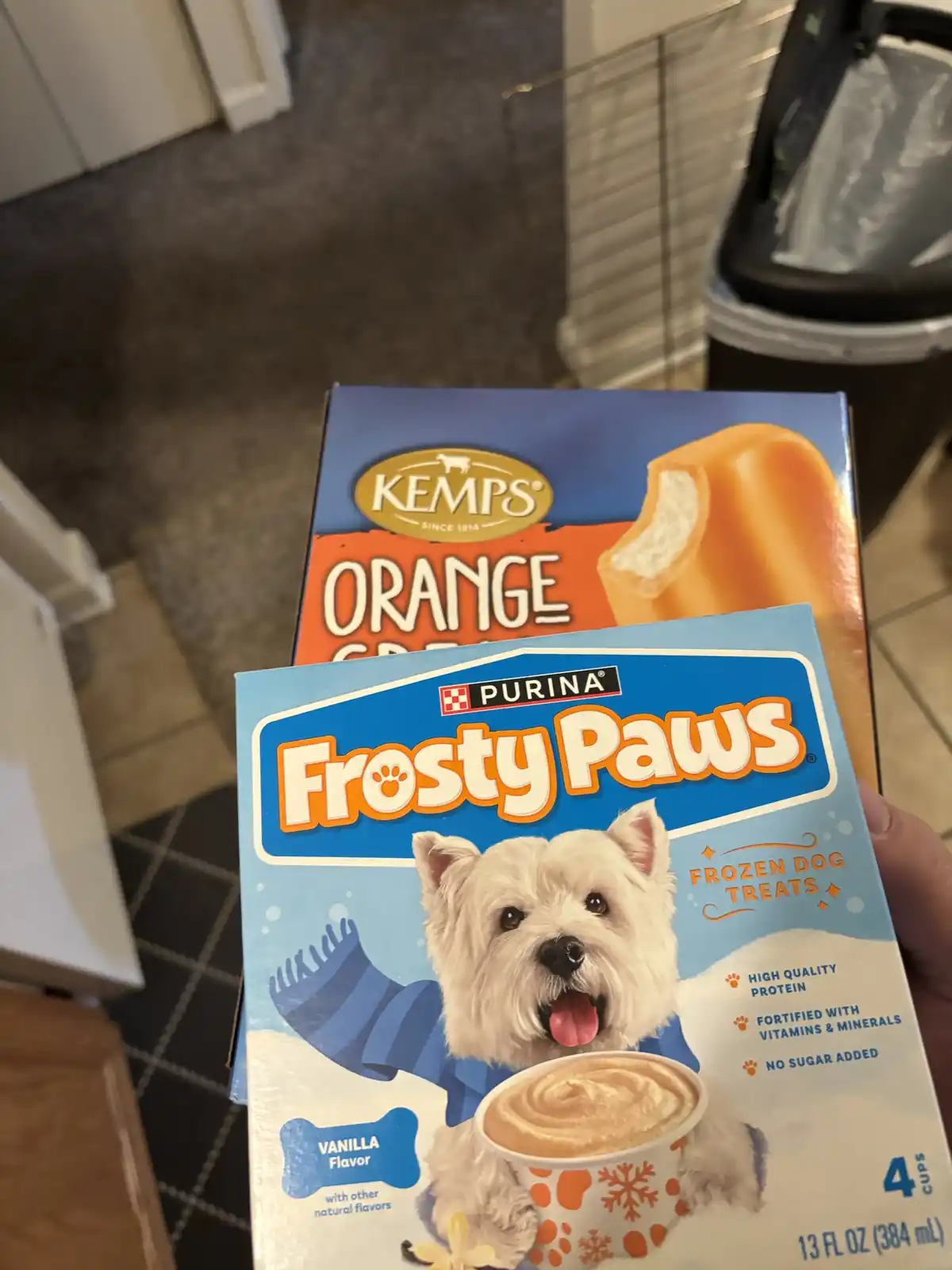 A hand holds a box of Purina Frosty Paws frozen dog treats in vanilla flavor, positioned in front of a box of Kemps orange…