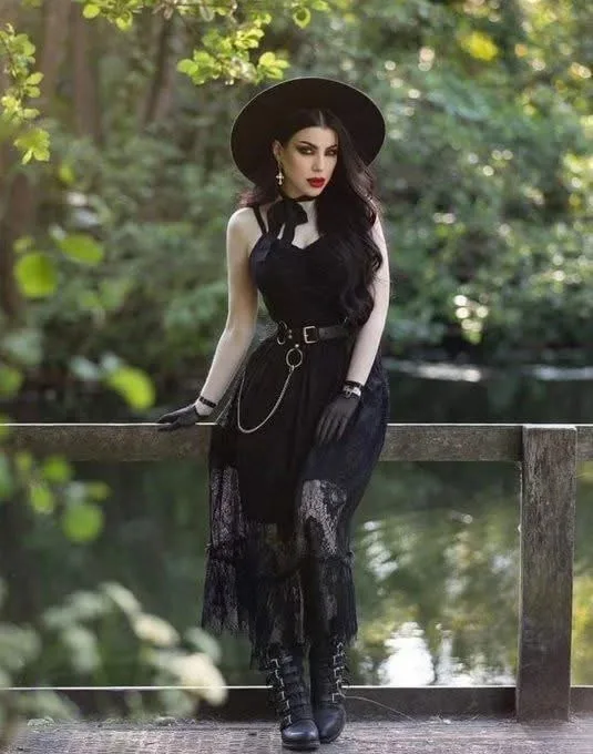 A woman in a full gothic witch outfit — black dress, wide-brimmed hat, lace, gloves, and boots — poses confidently on a…