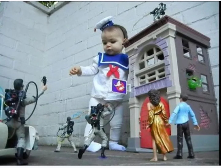 A baby dressed in a full Ghostbusters costume stands in the center of a miniature diorama featuring toy figures of…