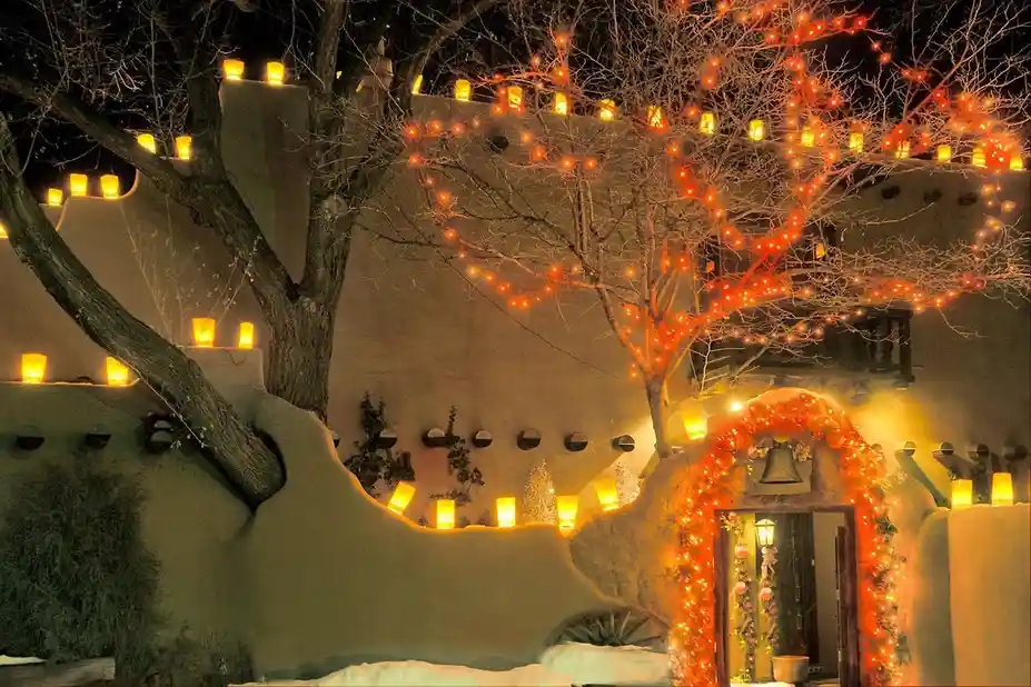 a nighttime photo of a snow-covered adobe-style building decorated with glowing lanterns and string lights on bare trees…