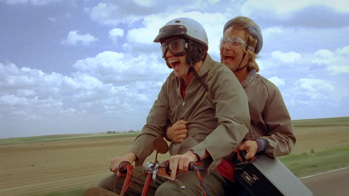 Two people wearing helmets and goggles are riding a motorcycle on a rural road, both laughing with wide-open mouths. The…