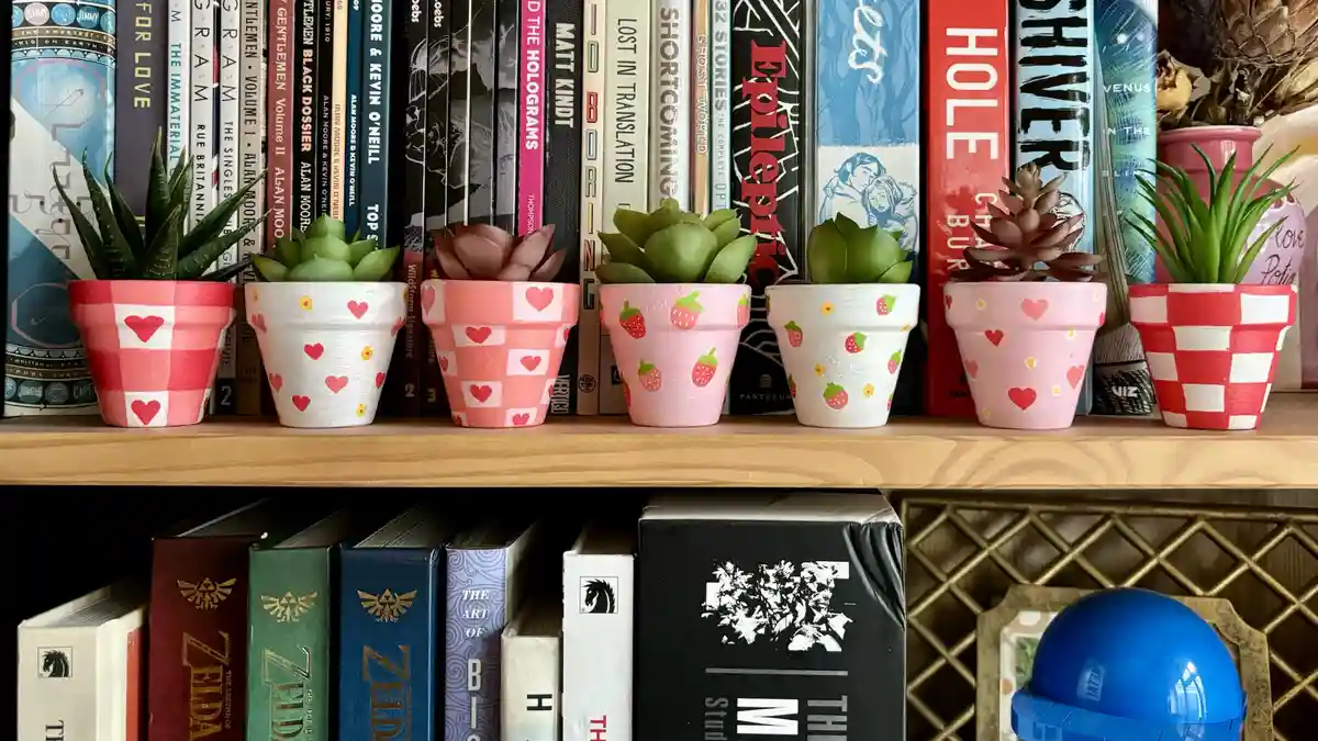 A row of six small, hand-painted terracotta pots with various pink and red patterns (hearts, strawberries, checkered) each…