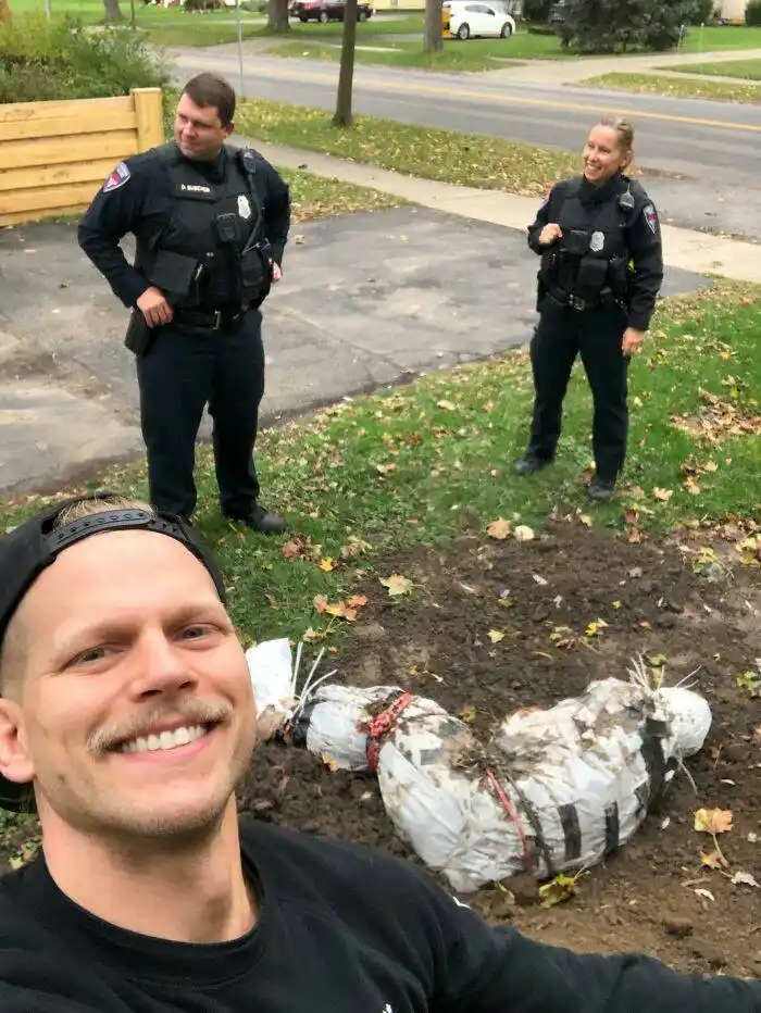 A man in a black cap and shirt takes a selfie smiling, with two police officers standing behind him in a residential yard…