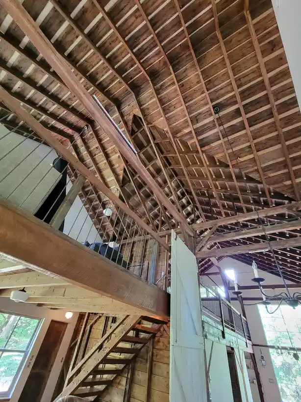A low-angle photo shows the exposed wooden beams and rafters of a high-ceilinged, rustic interior with a staircase and…