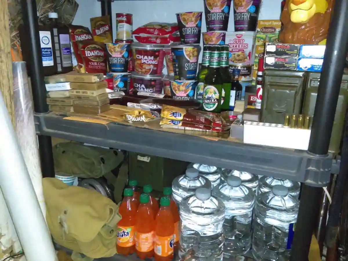 a cluttered metal shelf holds survivalist supplies including cup noodles, fanta soda, bottled water, ammunition, hydrogen…