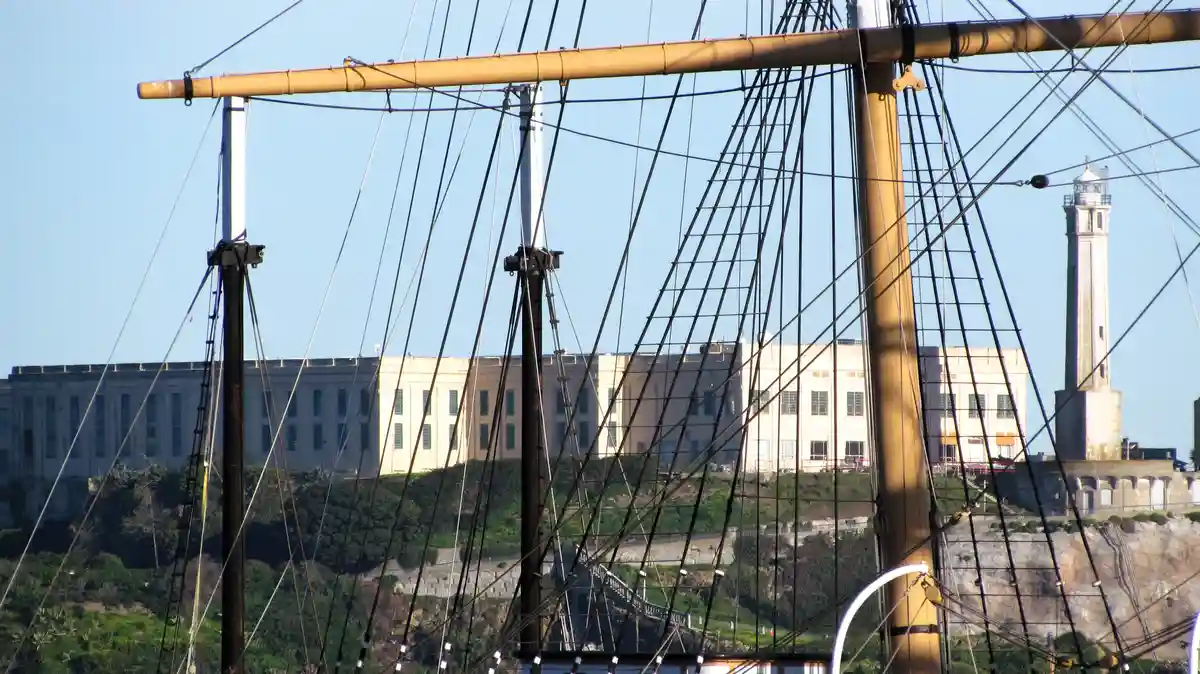 A view from a ship's rigging shows the historic Alcatraz Island prison and its lighthouse in the background, with masts and…