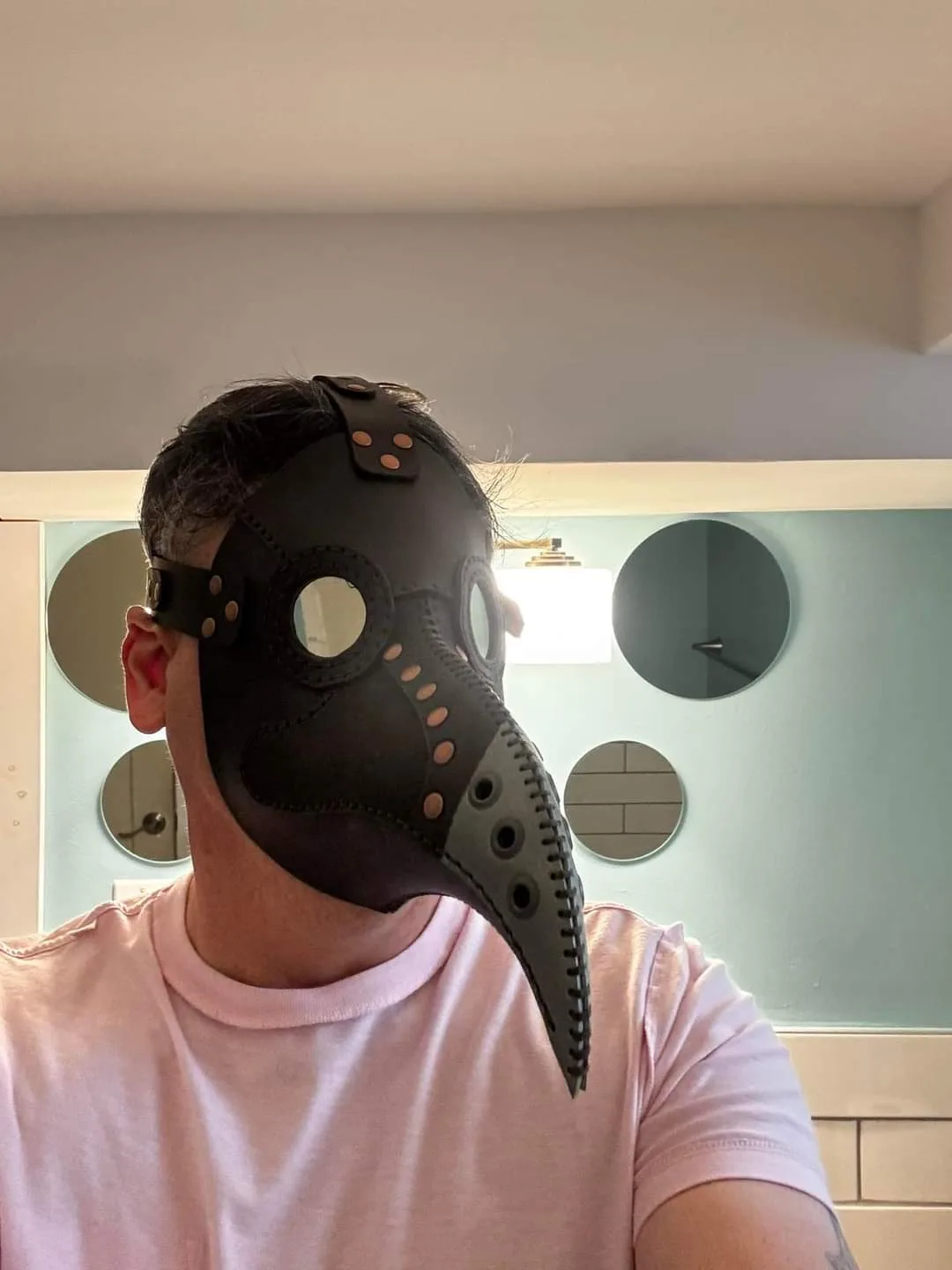 A person wearing a black leather plague doctor mask with a long beak and eye holes, standing in a bathroom with circular…
