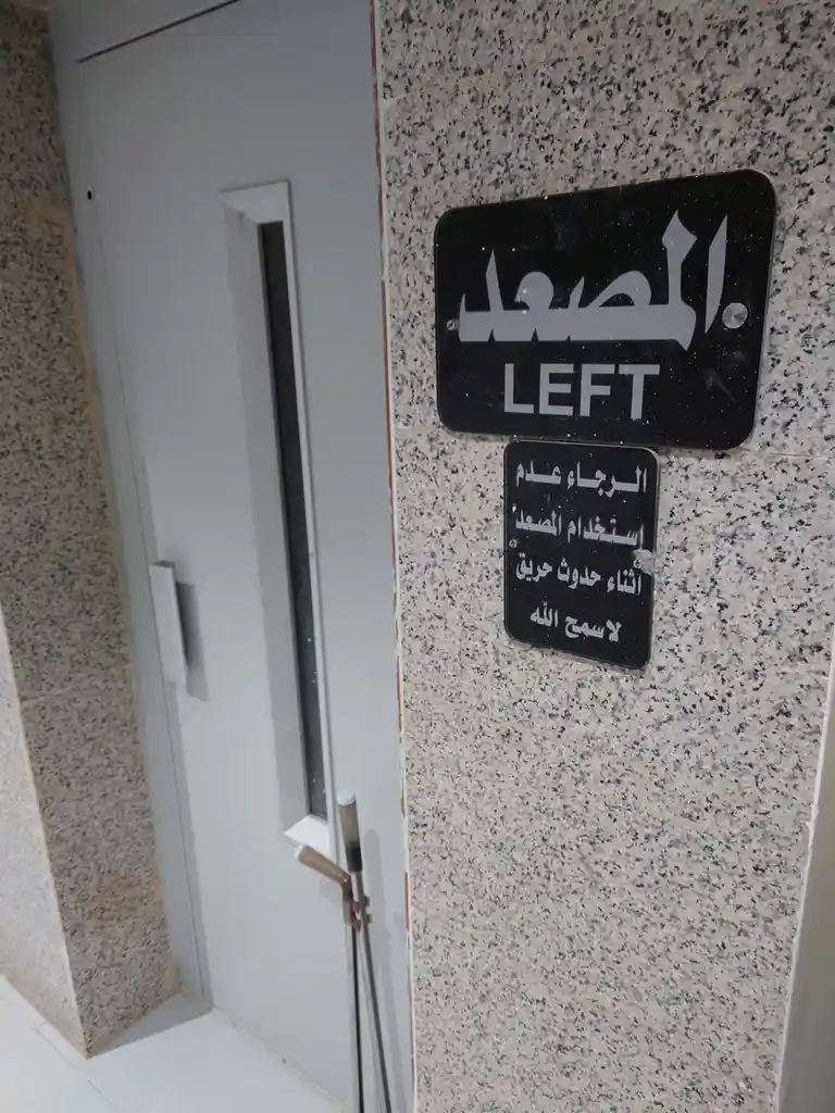 A photo shows a black sign next to an elevator door with Arabic text and the word 'LEFT' in English. The larger sign says…