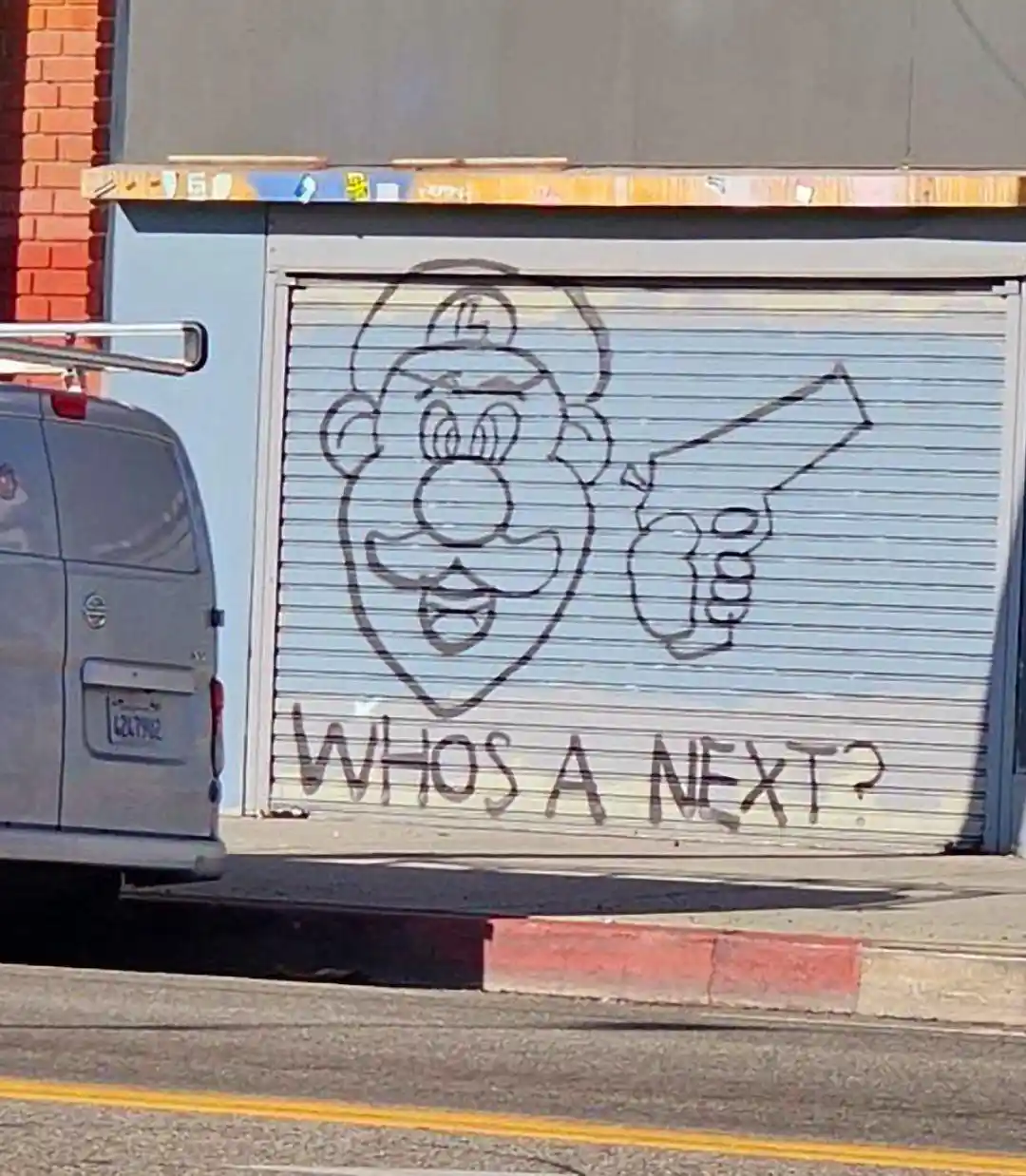 A light blue metal roll-up door has a crude black outline graffiti drawing of a cartoon character resembling Mario holding a…