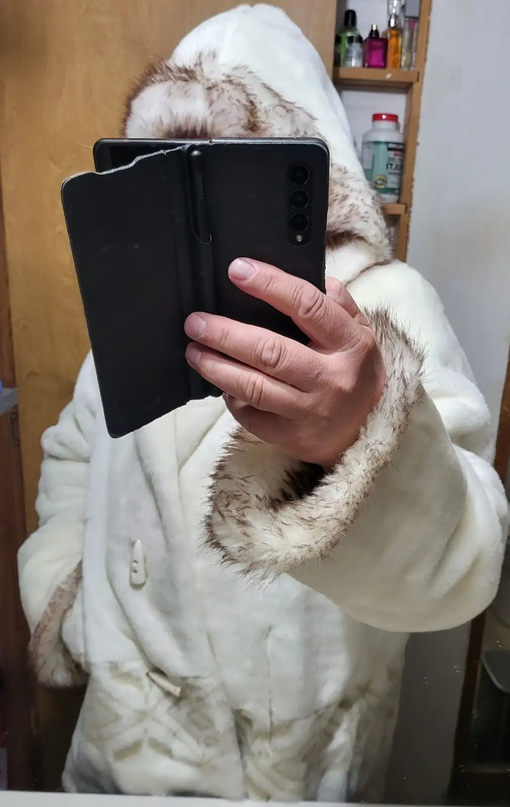 A person wearing a white, fluffy, fur-lined hooded robe holds a black smartphone with a case in front of their face…