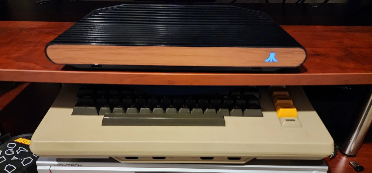 A black Atari 2600 console with a glowing blue Atari logo sits on a wooden shelf above a beige Atari 2600 keyboard with a…