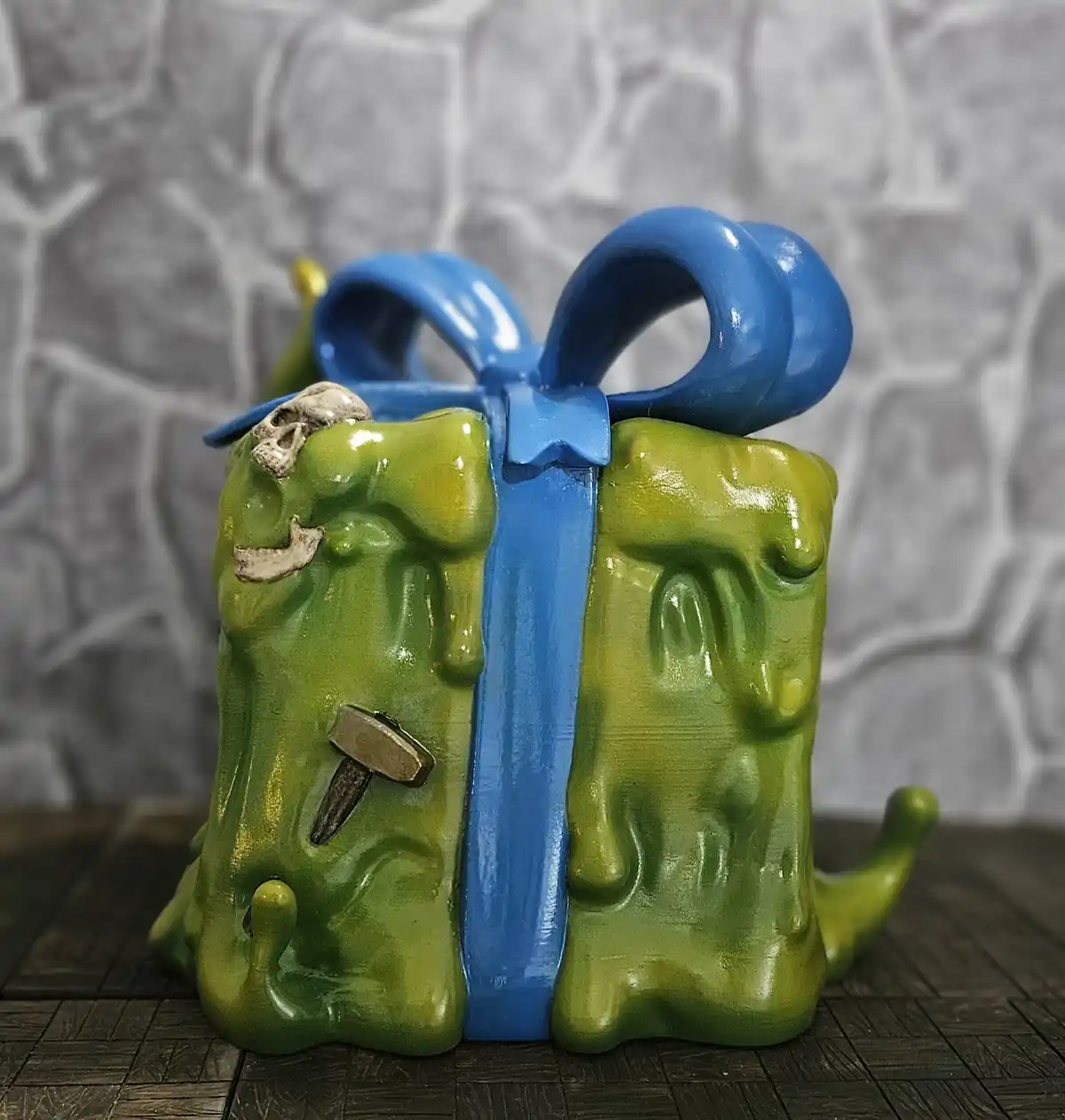 a green, grotesque, melting gift box with a blue ribbon bow, adorned with a small skull, a hammer, and other creepy…