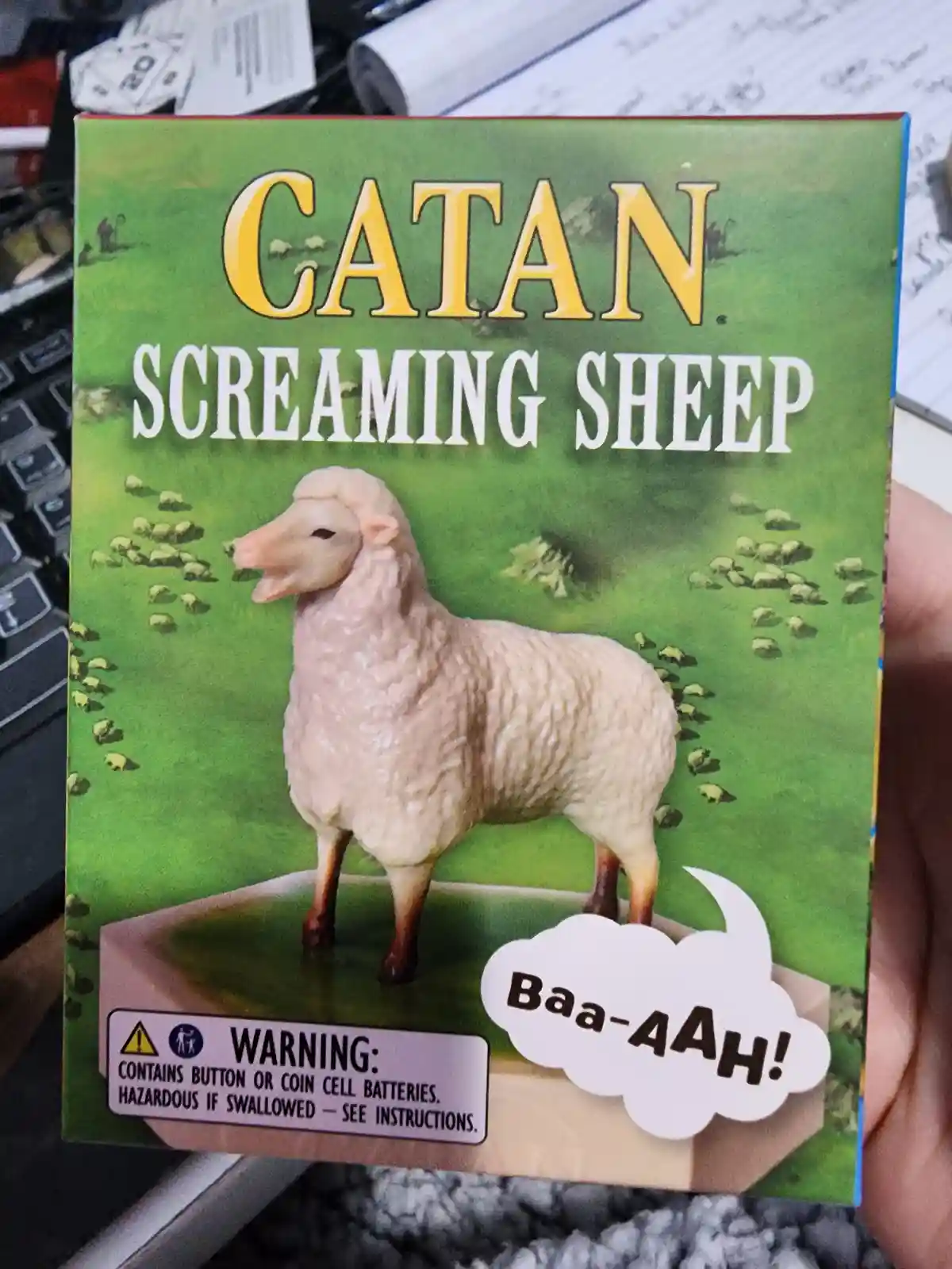 A person holds a box for a game called 'Catan Screaming Sheep', which features a plastic sheep figurine with its mouth open…