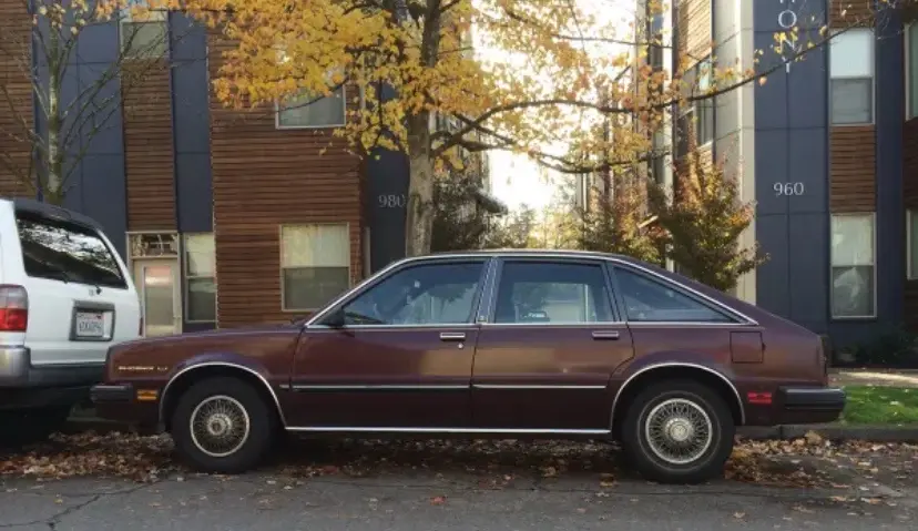 A maroon 1980s sedan is parked on a street in front of modern apartment buildings with autumn leaves on the ground. The car…
