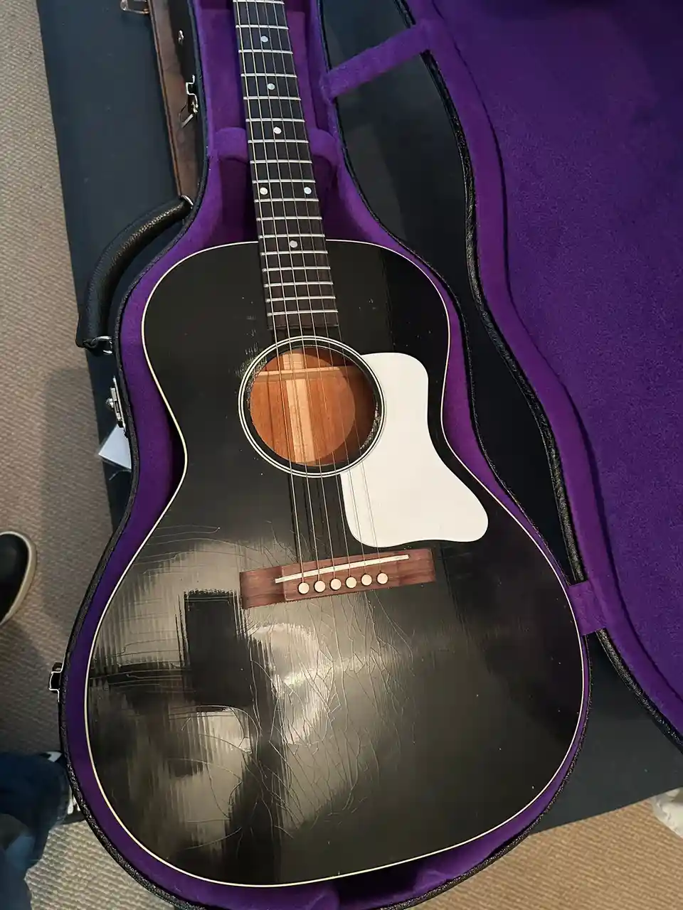 A black acoustic guitar with a white pickguard and visible scratches is resting inside an open purple case. The overlaid…