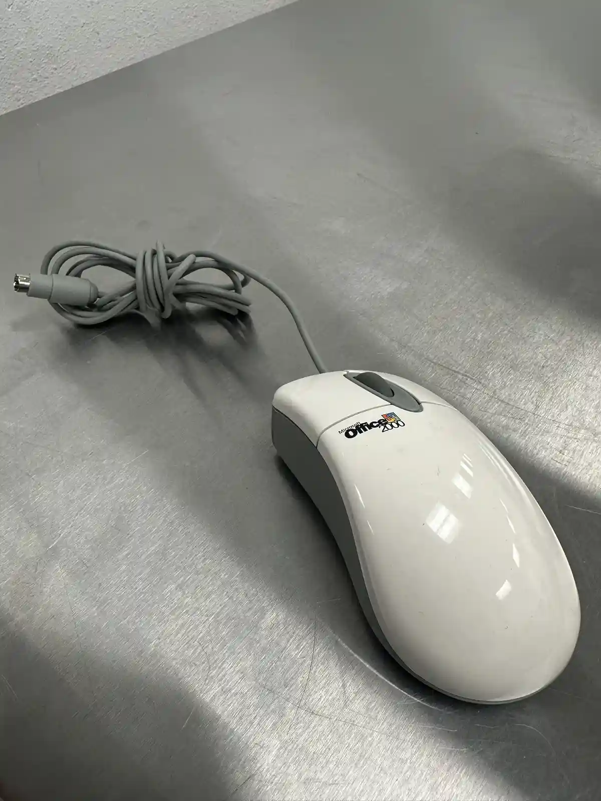 A white, wired Microsoft Office 2000 branded computer mouse rests on a scratched, metallic gray surface, with its gray cable…
