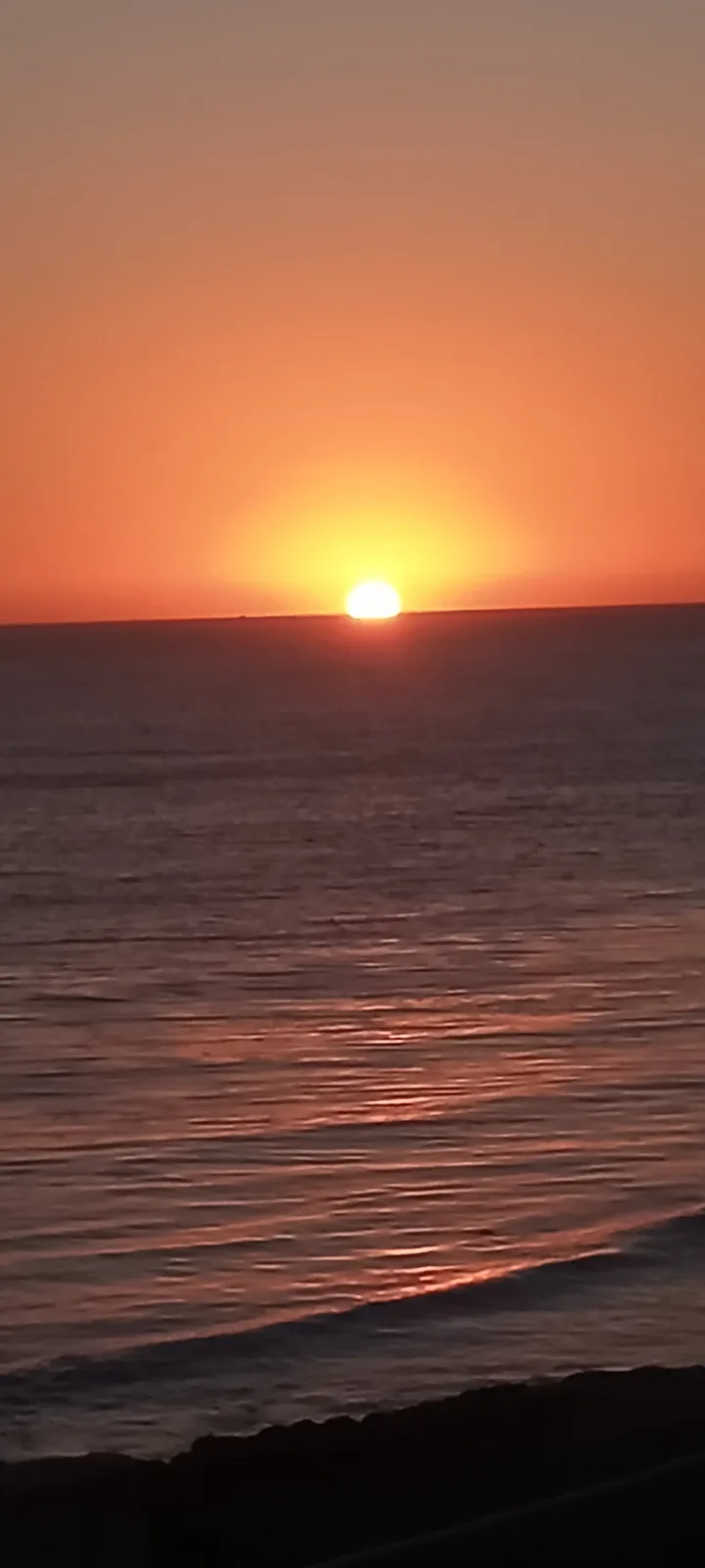 A serene sunset over the ocean with the sun low on the horizon, casting orange and red hues across the water and sky. The…
