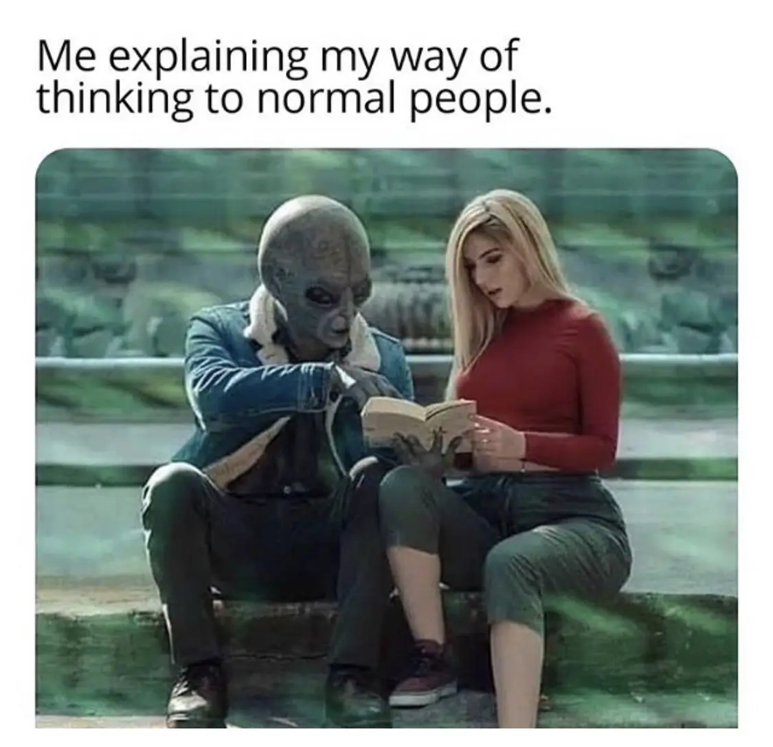An alien with a large head and dark skin sits beside a human woman in a red shirt, both looking at an open book. The alien…