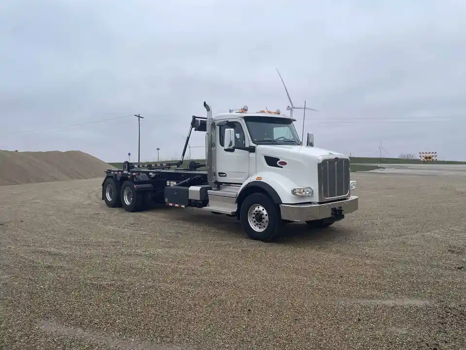 A white Peterbilt semi-truck with a flatbed and crane attachment is parked on gravel near a large pile of sand or gravel…