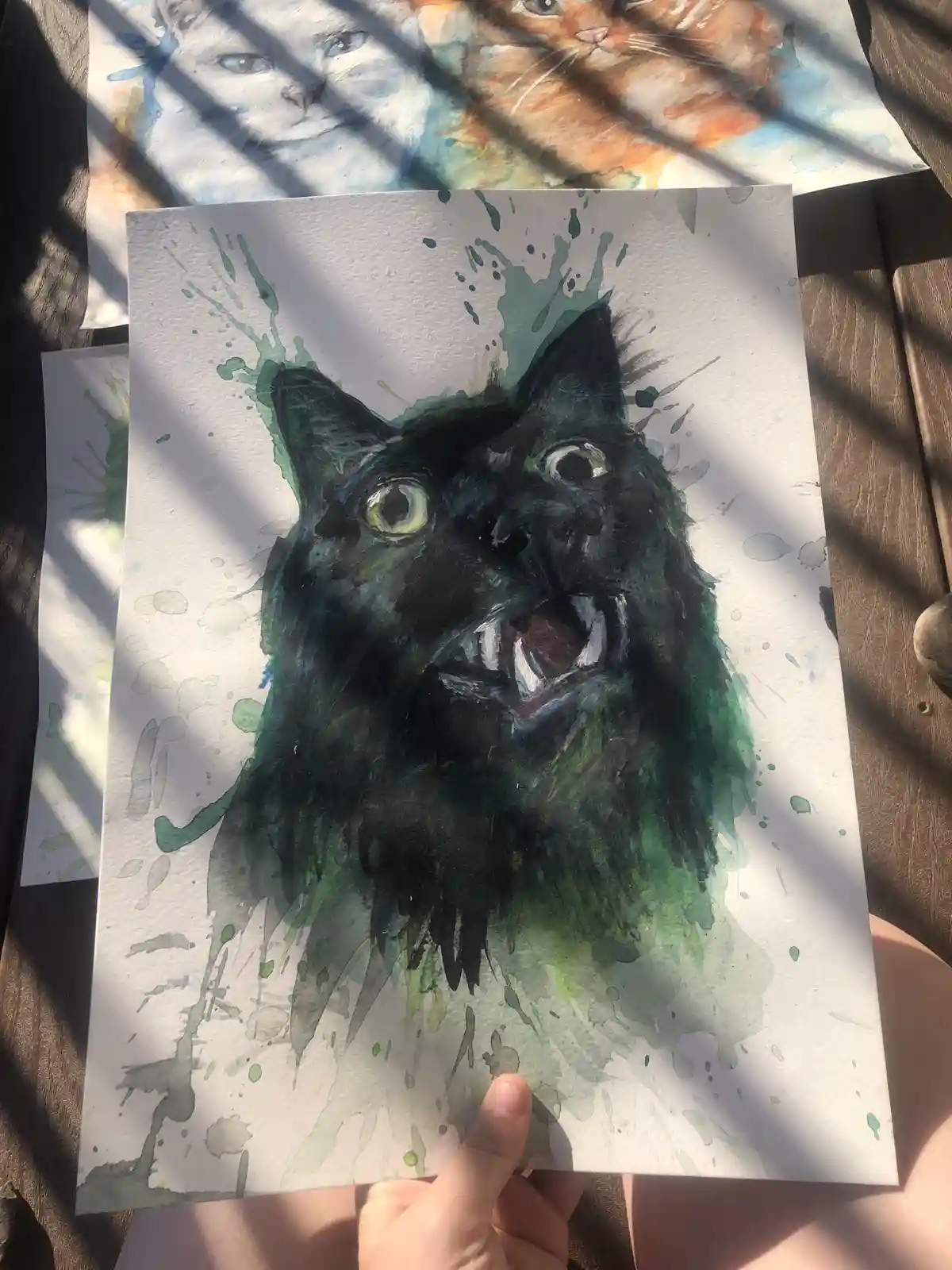 A hand holds up a watercolor painting of a black cat with wide, glowing eyes and bared fangs, surrounded by green splatters…