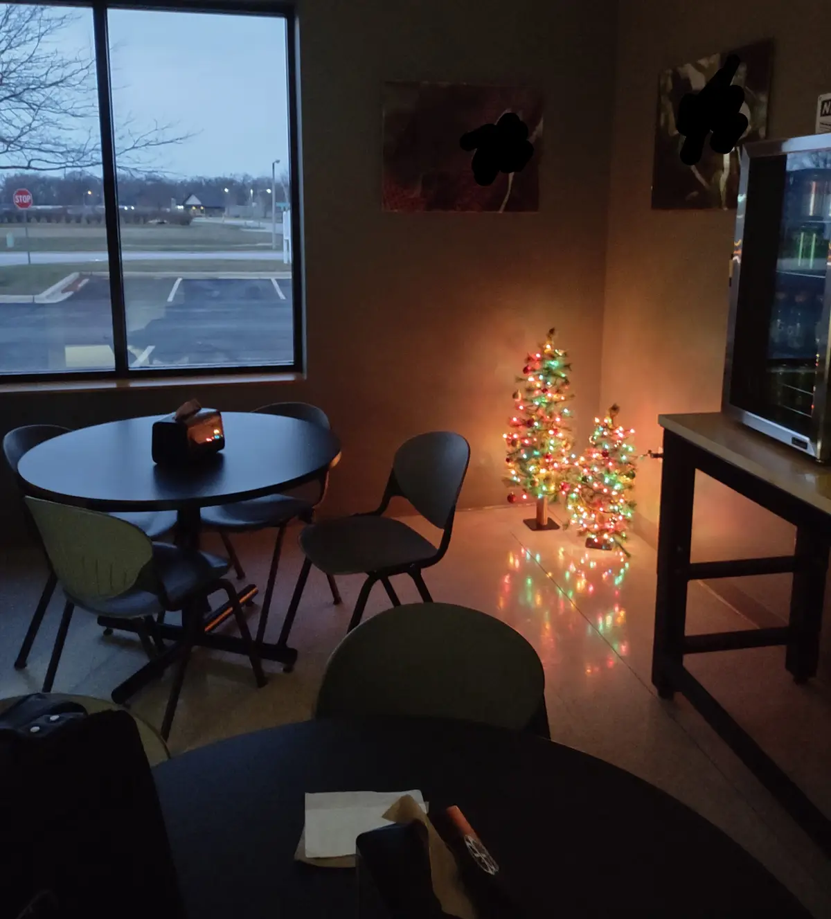 A dimly lit room with two round tables and chairs, featuring two small, brightly lit artificial Christmas trees in the…