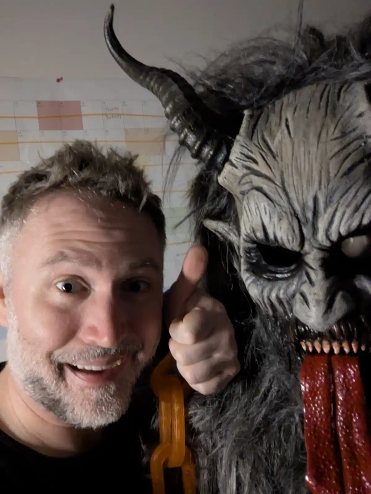 A man with a surprised, wide-eyed expression gives a thumbs-up next to a large, menacing demon mask with horns, sharp teeth…