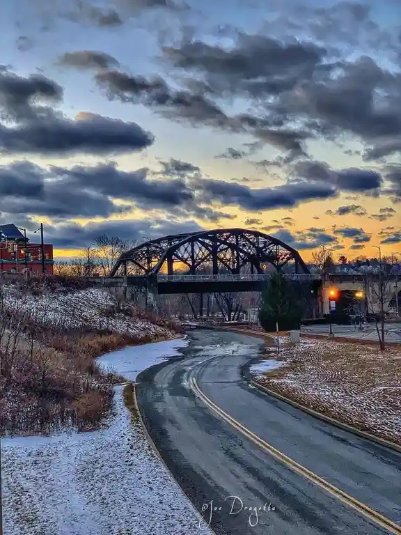 A scenic photo of a winding road leading to a metal truss bridge at dusk, with snow-dusted ground and a cloudy sky tinged…