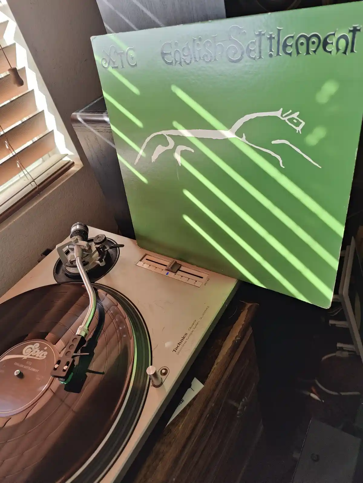 A close-up shot of a vintage Technics turntable with an XTC album titled 'English Settlement' playing, its green cover…