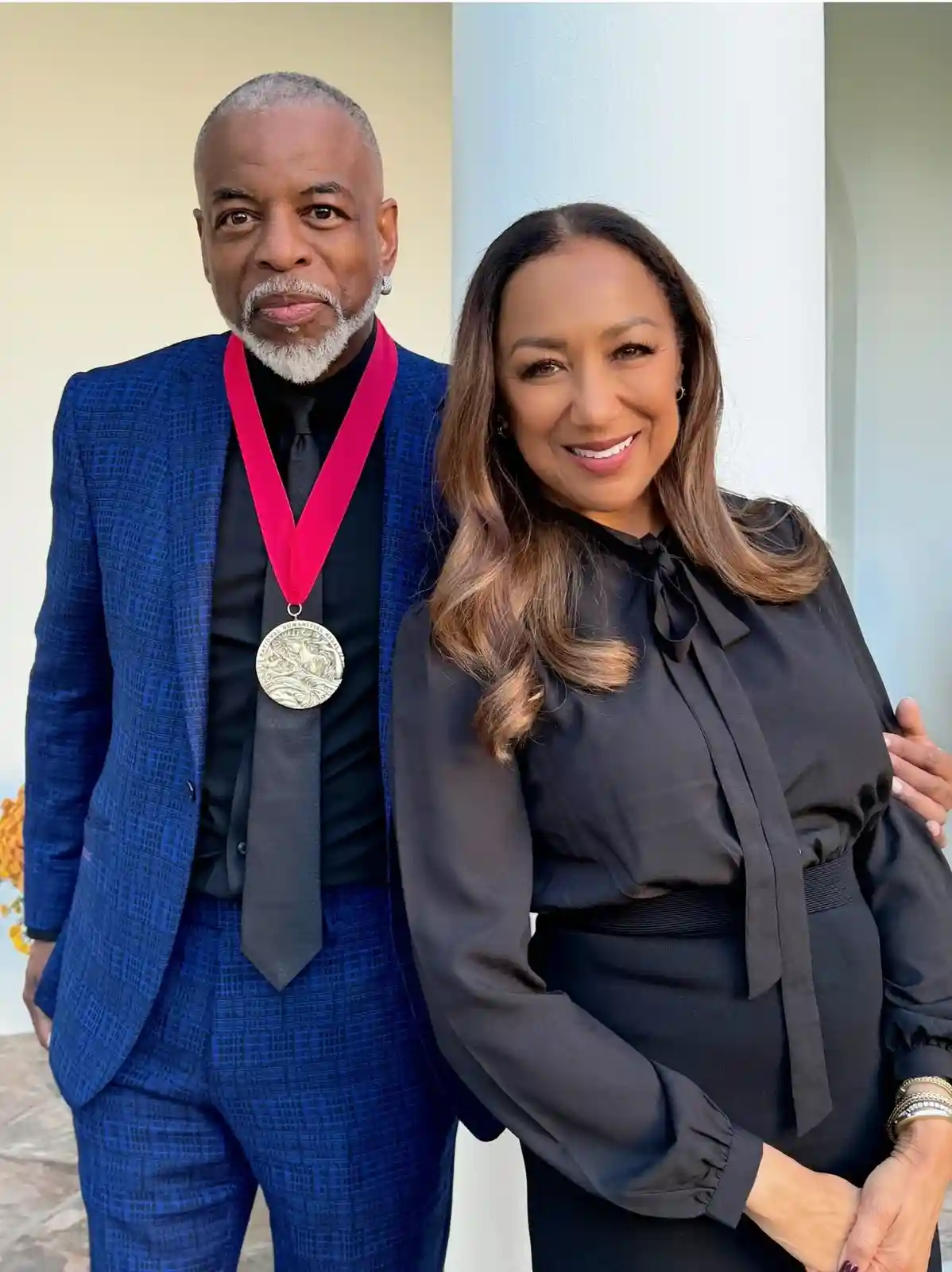 A man in a blue suit wears a large medal on a red ribbon while standing next to a smiling woman in a black dress. The man…