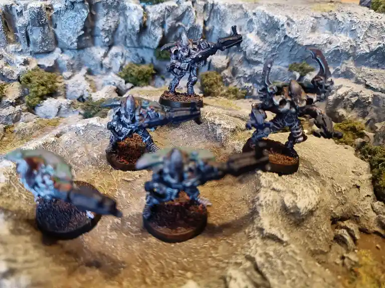 Five painted miniature figurines of grotesque, armored alien warriors with weapons stand on rocky terrain with mossy rocks…