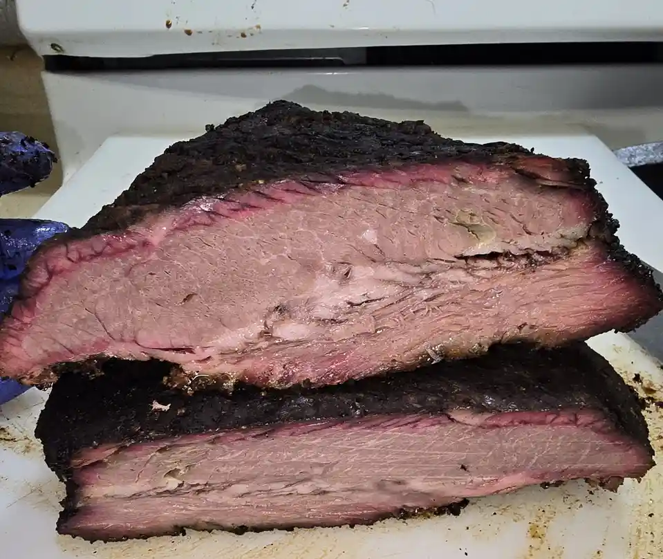 A close-up photo shows two thick slices of smoked brisket resting on a white cutting board, revealing a dark, charred…