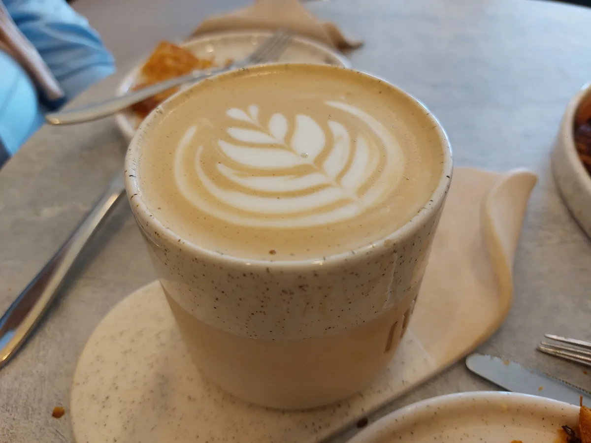 A close-up photo shows a speckled ceramic mug filled with a latte, featuring a white leaf-shaped latte art design on the…