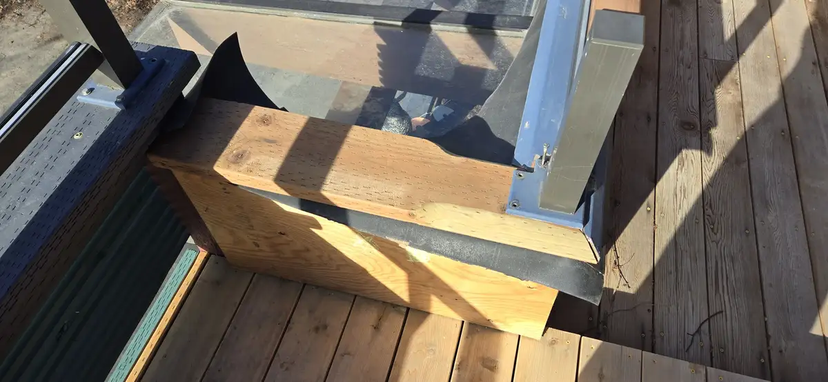 A close-up photo shows a wooden deck railing with a freshly installed wooden beam and metal brackets, set against a wooden…