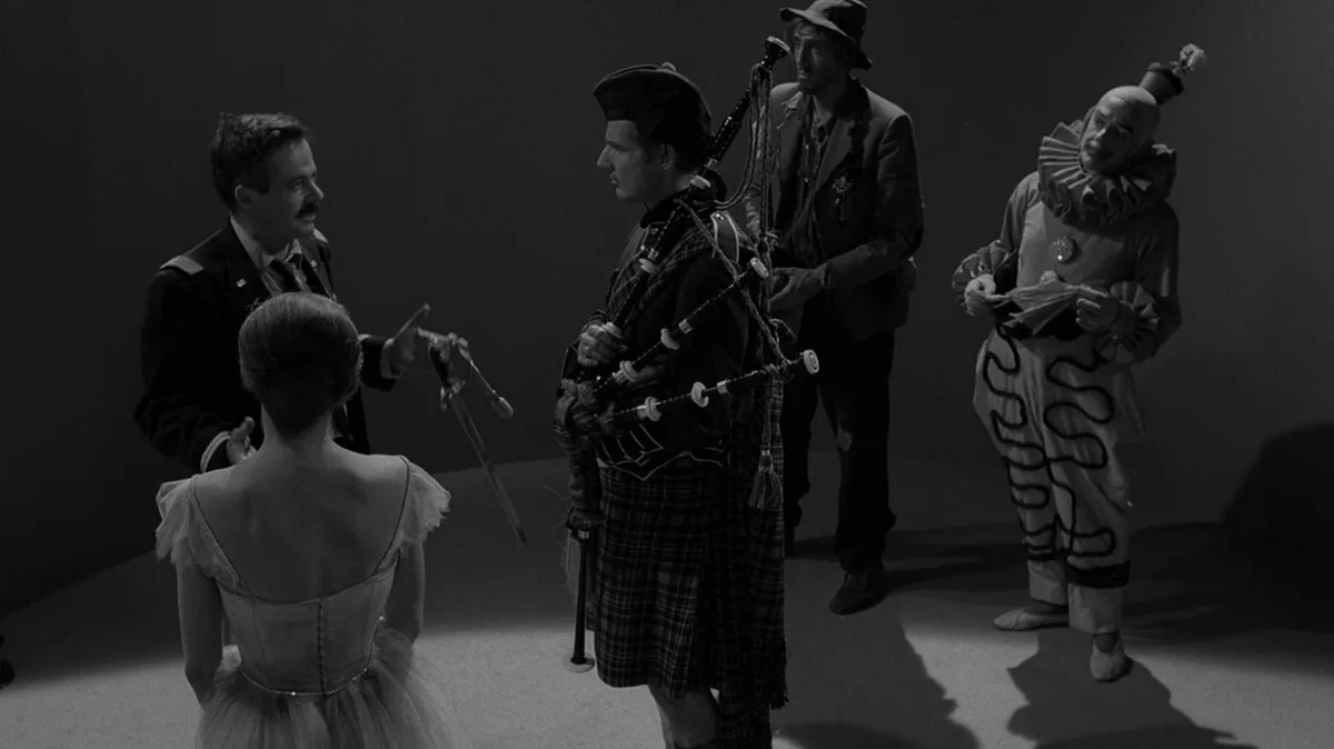 A black-and-white scene from a film shows a man in a military-style uniform gesturing toward a ballerina in a tutu, while a…