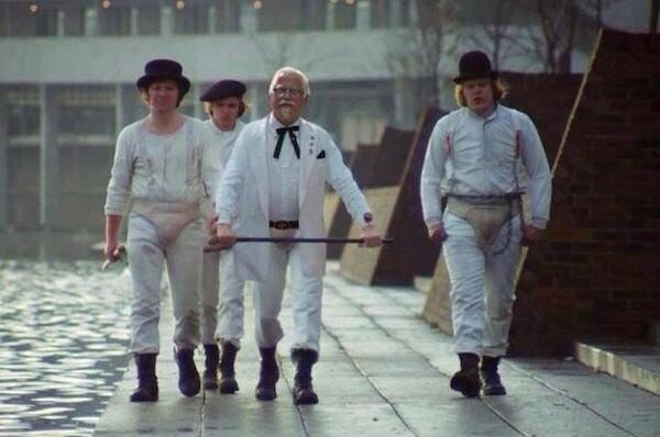 Four men in white outfits and bowler hats walk in a line, led by an older man in a white suit holding a long stick. The…