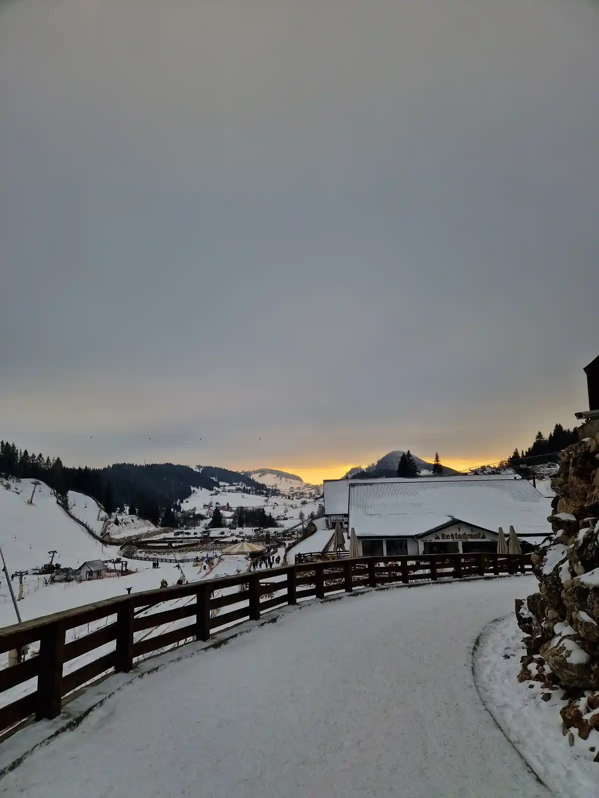 A snowy mountain resort scene at dusk, featuring a winding snow-covered path with a wooden railing, a building labeled…